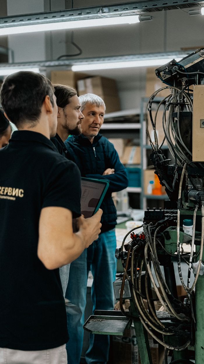 why-choose-us-02 Three adults engaged in a discussion around complex machinery in a factory environment.